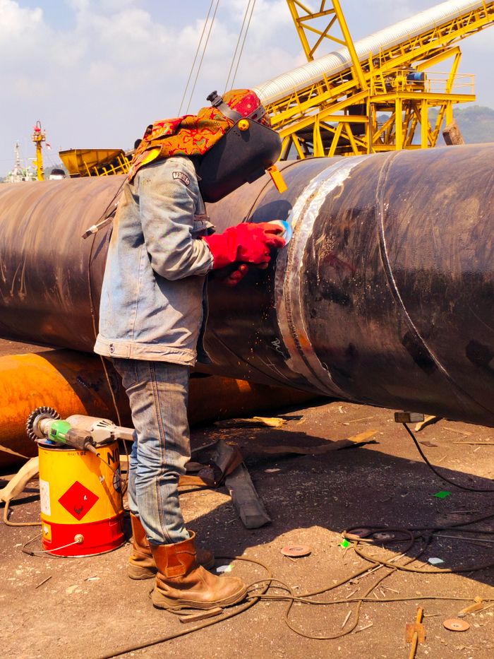 A welder in protective gear welds a large metal pipe outdoors on a sunny day.