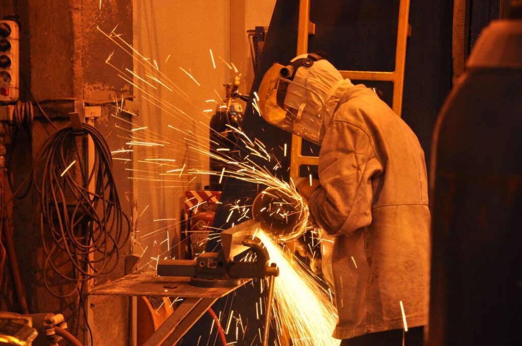 A worker in protective gear uses a grinder in an industrial setting. Sparks fly in the indoor workshop.